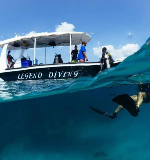 Diver with fins swims underwater beside a boat labeled Legend Diving as crew prepare gear off Nusa Lembongan.
